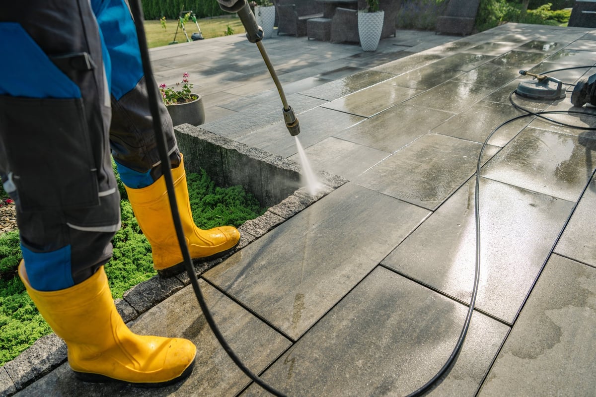 A worker in yellow boots cleaning a stone patio with a pressure washer on a sunny day